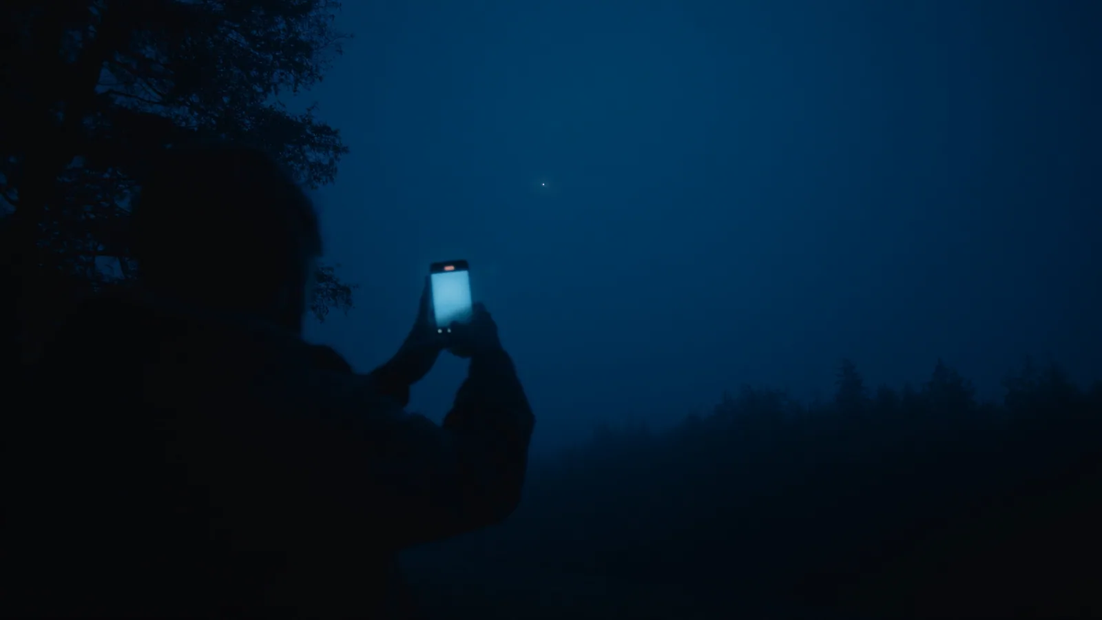Silhouetted hands holding a phone aimed at a deep blue night sky, a single bright unidentified light visible above the treeline.